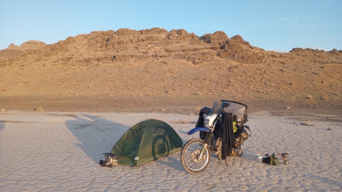 A DR650 Suzuki motorcycle is parked next to a 1-person tent on a cracked dry lakebed. It is sunrise and the front wheel's shadow sita on the tent. Behind is a lrge outcrop of rocks.