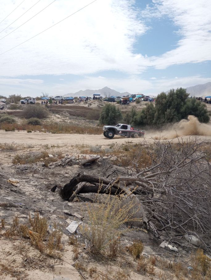 Trophy truck slams through a deep rut in a dry creek bed, front wheels in the air and rear wheels fully compressed, with hundreds of spectators and canopies lining the bank of the creekbed in the background. In the foreground, a dead and burned out tree.