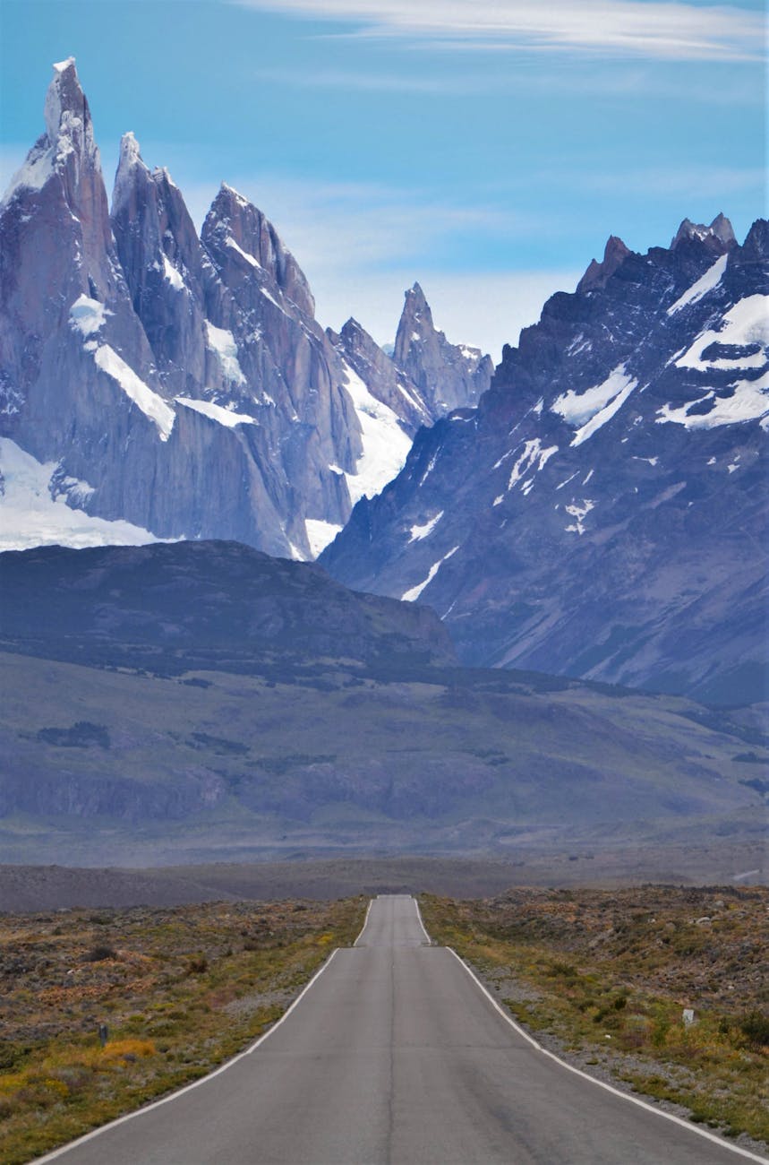 A long, straight band of asphalt with no center line wanders into the distance. Sharp peaks are further out, covered in snow. The Andes mountain range most likely.