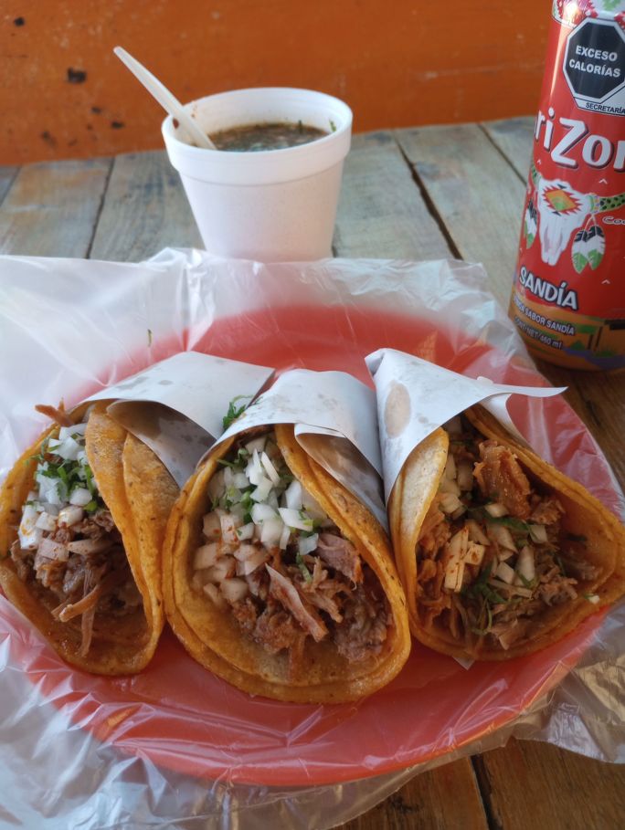 three birria tacos on a plate, wrapped in paper, with onions and cilantro. Behind them is a cup of consome, and a can of fruit punch, all on a wooden, outdoor table