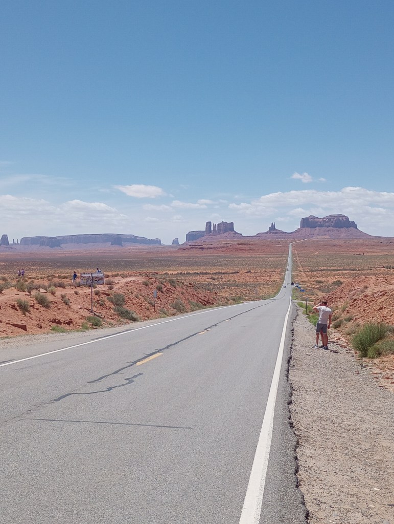 Monument Valley in Utah, at the Forrest Gump vista. Desert sand in browns and reds mix with a few sage brush while a straight two lane highway cuts through a wide valley to the spires and mesas in the distance.