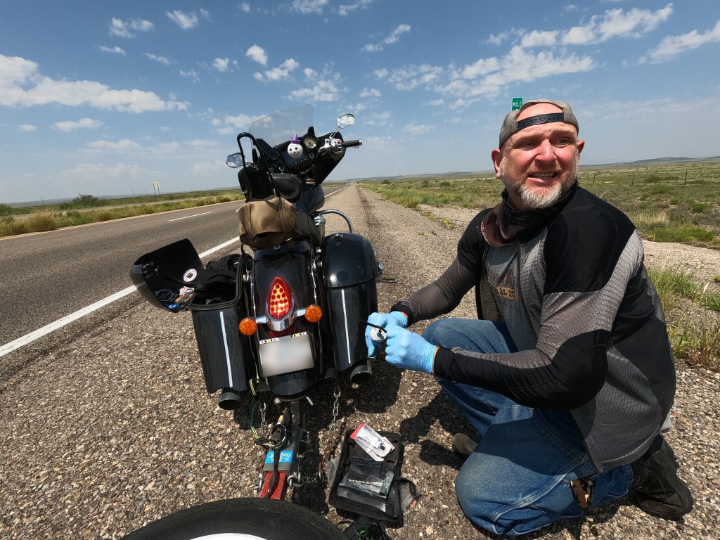 Man on side of highway repairing flat tire on motorcycle. He looks back toward the camera, and to approaching traffic to verify safety