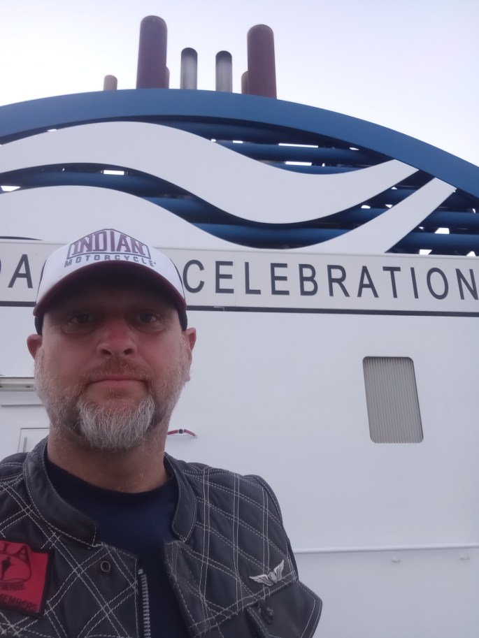 Person standing on a ferry looking at the camera. Behind, on the ship's superstructure, is painted "celebration."