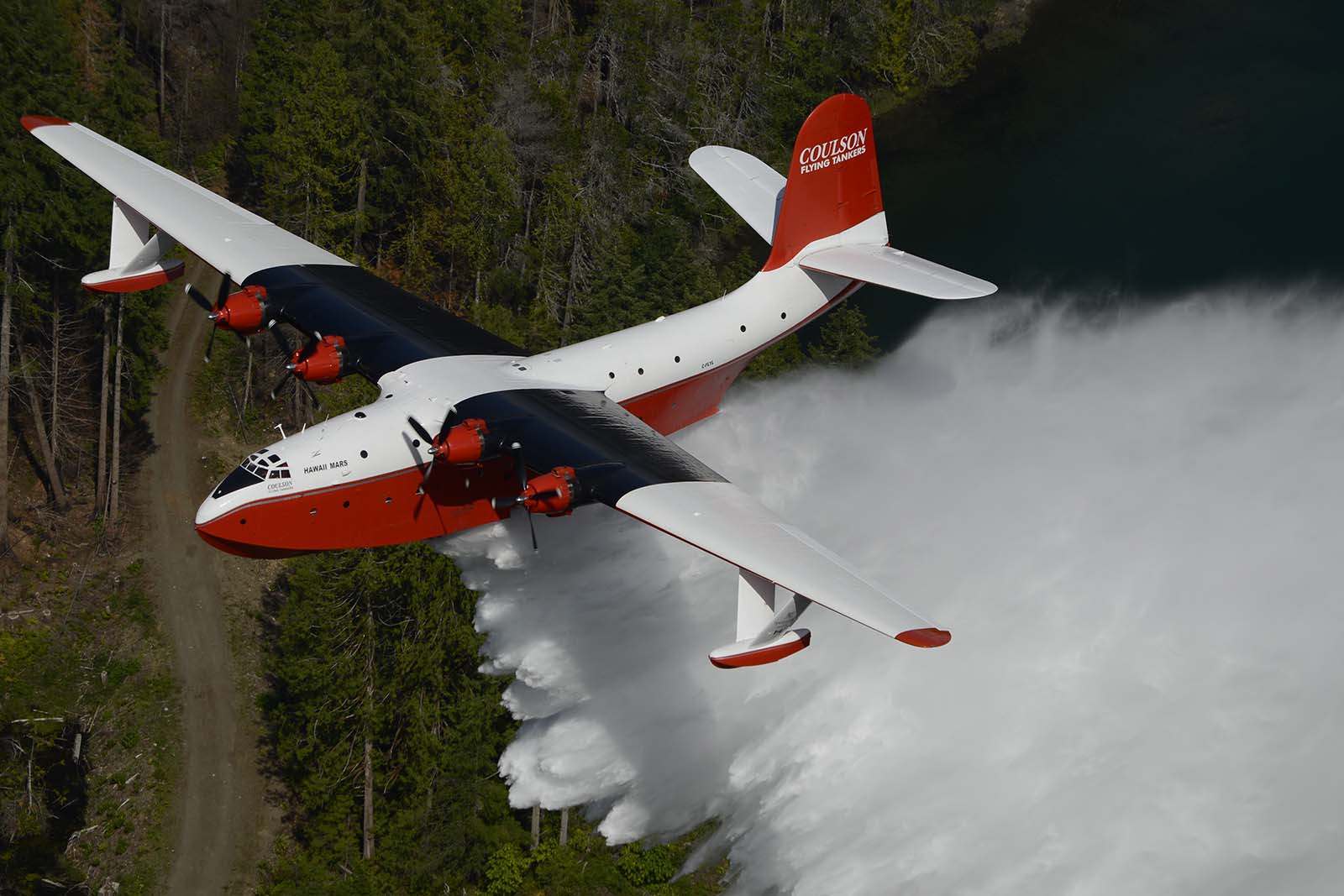 Martin Mars water bomber in action, dropping 7,000 gallons of water. Viewed from above and at the left-front quarter, the flying boat is white on top with orange-red lower half. Its wings are painted black behind its engines to hide exhaust smoke stains. The tail reads "Coulson" and below the huge plume of water is nearby forest, service road, and the edge of a lake