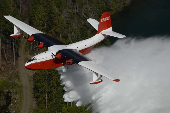 Martin Mars water bomber in action, dropping 7,000 gallons of water. Viewed from above and at the left-front quarter, the flying boat is white on top with orange-red lower half. Its wings are painted black behind its engines to hide exhaust smoke stains. The tail reads "Coulson" and below the huge plume of water is nearby forest, service road, and the edge of a lake