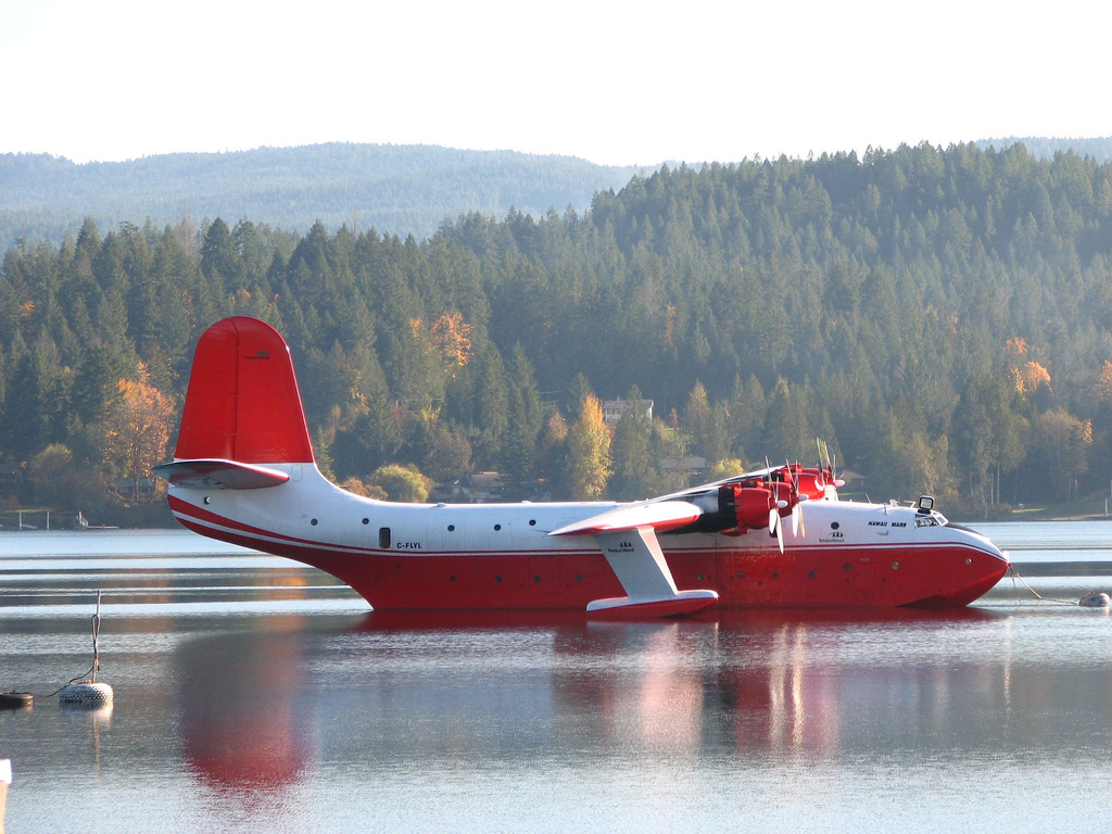 JRM Hawaii Mars water bomber, a massive four engine flying boat from the 1940's, rests on Sproat Lake on Vancouver Island, thethered to its mooring. In the backgournd are low mountains with trees, some chaging color for the fall. The water is placid