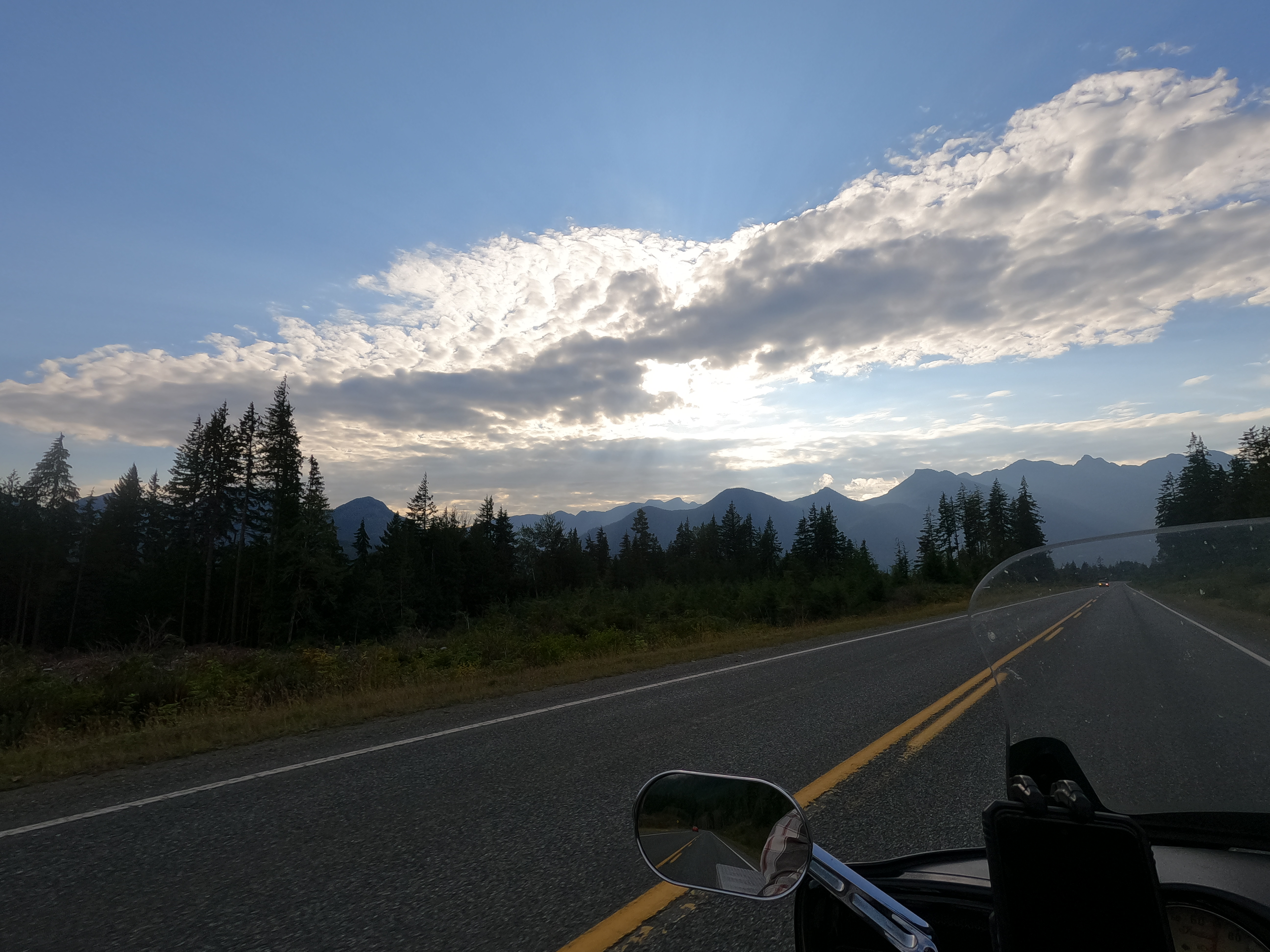 POV of a motorcyclist on a forest highway, looking out at tall trees and scattered clouds with the sun shining behind them. Firther on the horizon are gray mountain peaks