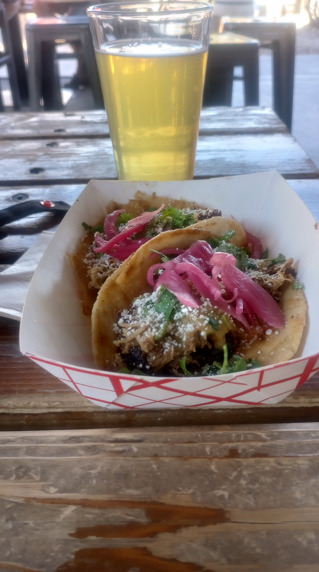 Two nice looking but overpriced tacos sit in a paper basket with a pint of beer in a glass in background, sitting on wooden outdoor bench
