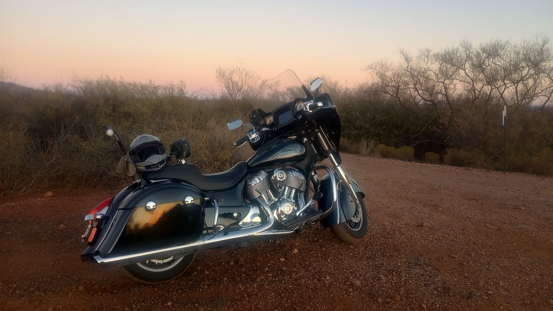Indian Chieftain motorcycle black, parked on dirt road in desert setting with low trees, sage, and sunset