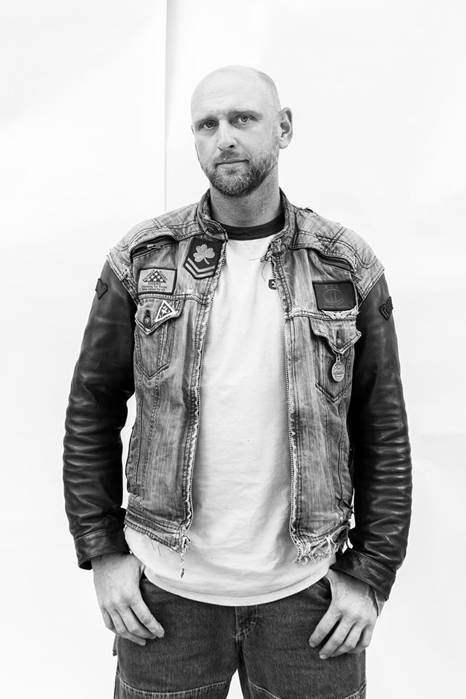 grayscale portrait of Johnny Killmore standing wearing motorcycle jacket jenim and leather with patches, light t-shirt, and blue jeans