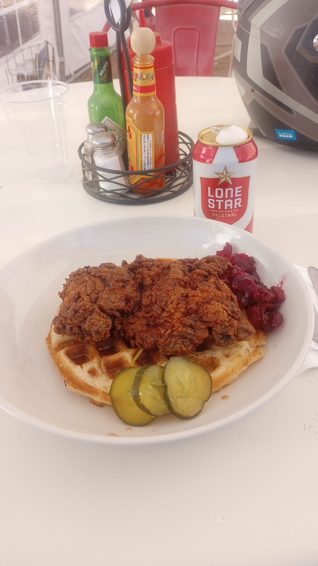 a plate of chicken and waffles, with cranberry sauce and dill pickles as garnish, sits on an outdoor restaurant table with a Lone Star beer can and condiment caddy