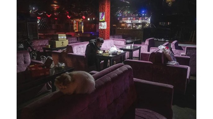 A center-lit scene of a man sitting inside an empty table at a bar and restaurant, eating a simple meal. A cat sits in the foreground on a seatback, boxes and bags sit waiting to be unpacked, and a motorcycle is parked in an aisle.