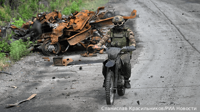 Soldier with tactical gear and goggles rides a muddy dirt bike on a road, passing rusted and long destroyed vehicle remains