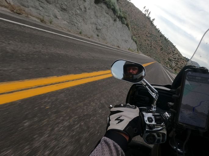 POV of a motorcyclist leaning into a corner on a mountain road. The rider's face reflects back at the camera from the mirror as he stares through the corner, intently reading the road ahead.