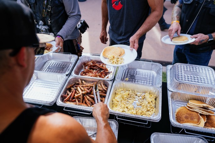 volunteers serve breakfast in an outdoor setting. Sausage, bacon, eggs, and pancakes are ready as a plate is filled