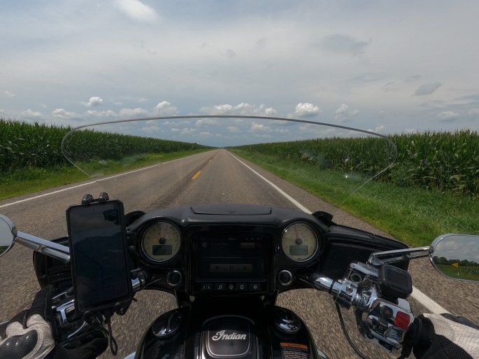 POV of a motorcyclist on a two lane road in middle America, with tall cornfields on either side. The sky has a slight haze of overcast and low puffy clouds in the distance