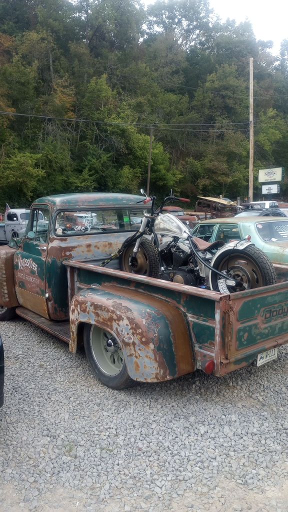 A custom bobber style motorcycle in the bed of a 1950's Ford pick up, which has faded paint of forest green mizing with rust and primer.