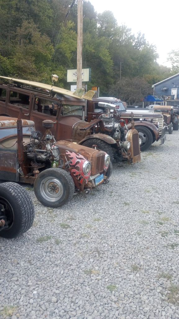 A line of rat rod customs of varying style, from large trucks to 1930's cars with 1950's grill work, parked in a gravel lot at a car show