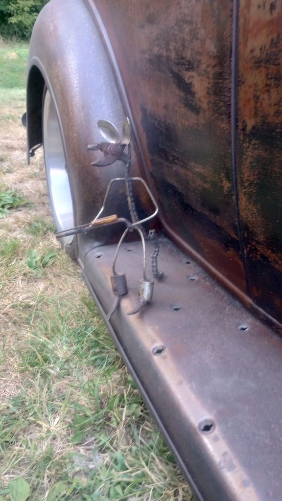 a tiny figure made of bet steel and small bolts is welded to the running board of an old car. It has the shape of a humanoid mouse holding a machine gun