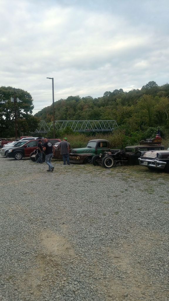 Old clay's ferry bridge across the kentucky rive in the background. in the foreground are rat rod custom cars with a few 1950's American cars and a few modern cars further behind, in a gravel lot with green full trees all around.