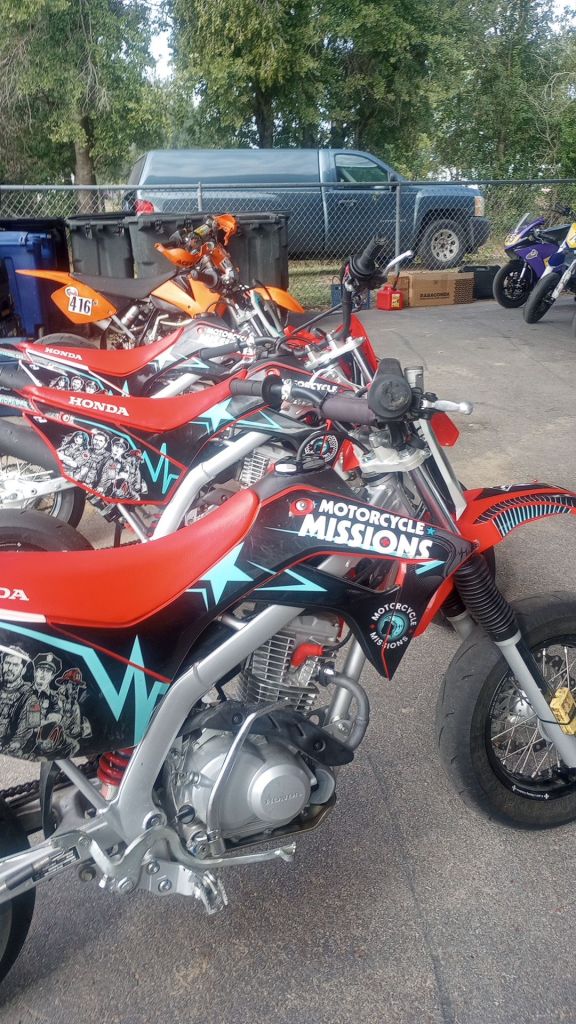 Motorcycle Missions branded motorcycles sit lines up in a racing paddock.