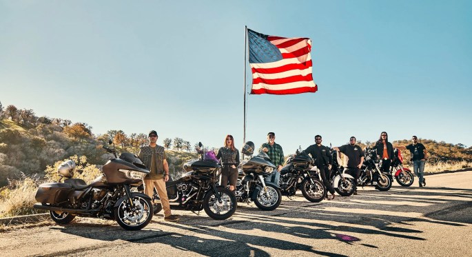 Harleys and their owners post on the edge of a road with a large American flag in the background