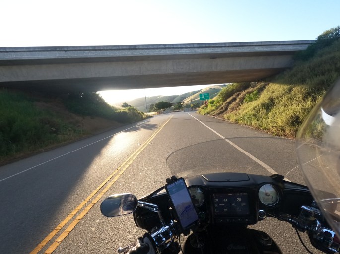 POV of a motorcyclist riding into the sunset on a 2-lane road, passing under a highway bridge.