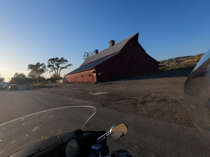 POV from a motorcycle, looking over at a large red barn with a peaked roof on the side of the road