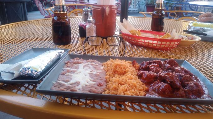 Chile Colorado on a plate with rice and beans. In the background is a bottle of beer, basket of tortilla chips, and salt and pepper shakers.