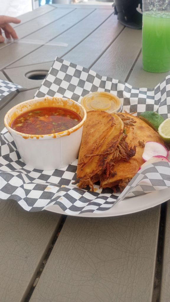 Birria tacos in a basket with consome, salsa, radishes and lime slices, on a plate with a checkerboard pattered paper