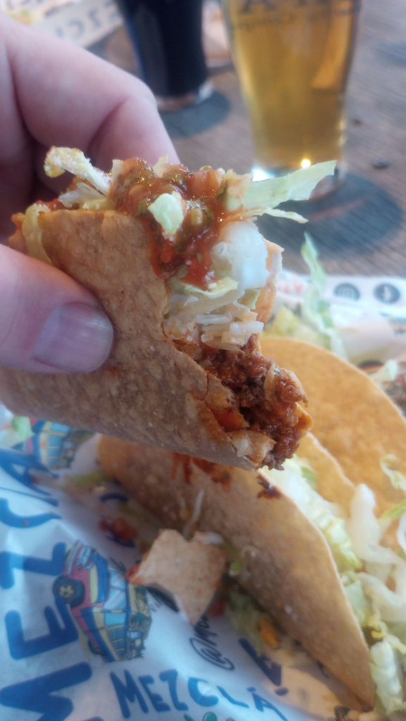 a half eaten hard shell taco in hand, with other tacos in the background.
