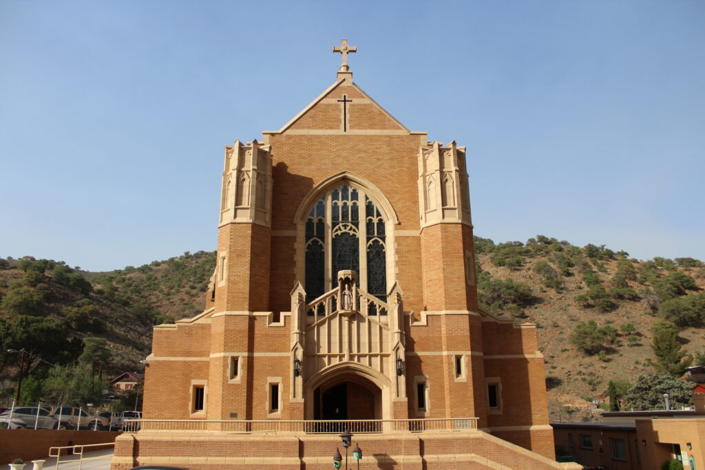 the front of a catholic church with spires and cross, tall glass window, and statue of saint patrick, with desert hillside and greenery rising up in the background, meeting a clear sky