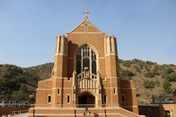 the front of a catholic church with spires and cross, tall glass window, and statue of saint patrick, with desert hillside and greenery rising up in the background, meeting a clear sky